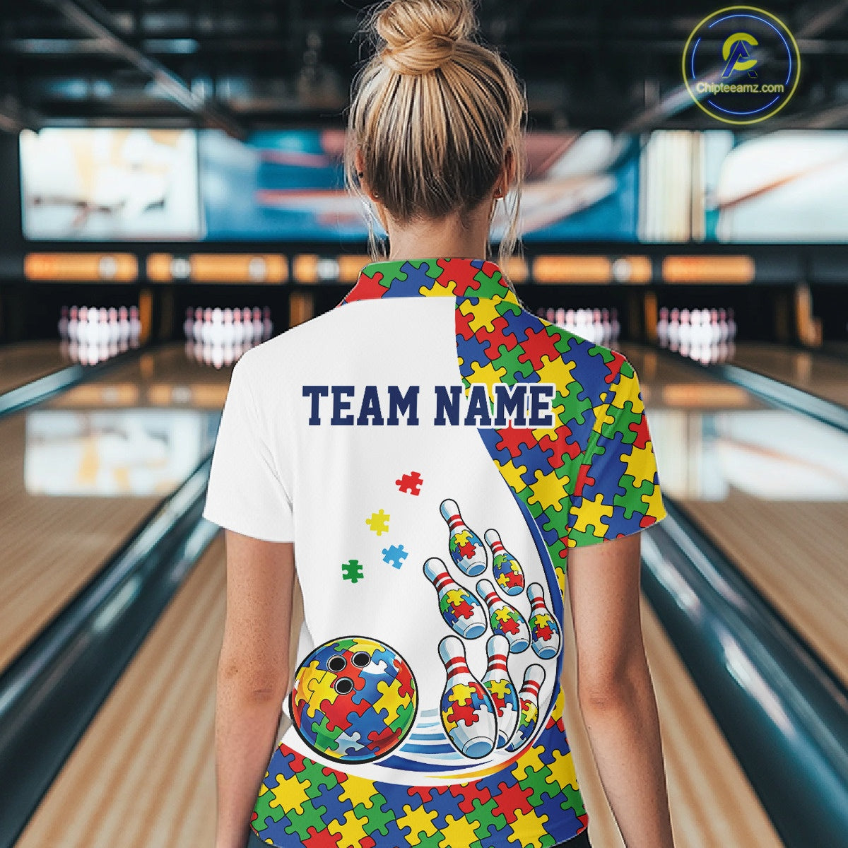 Back view of a female bowler wearing a white quarter-zip shirt, colorful autism puzzle-themed bowling design with dynamic strike elements.