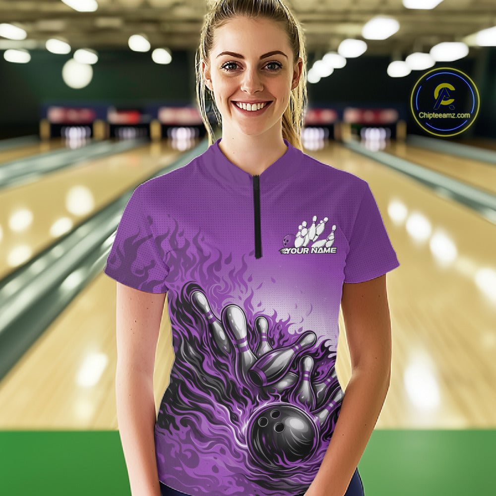 In a bowling lane, female model wearing a purple quarter-zip shirt, flame bowling ball striking pins with vibrant fire-inspired sports graphics.