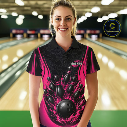 A woman stands in a bowling alley wearing a black and pink short-sleeve polo shirt with a bowling pin and ball graphic on the front. 