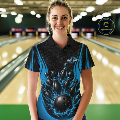 A woman stands in a bowling alley wearing a short-sleeve polo shirt with a black and blue bowling pin and ball graphic on the front. The shirt has a black collar and shoulders, bright blue flame-like side panels, and a dark patterned center.