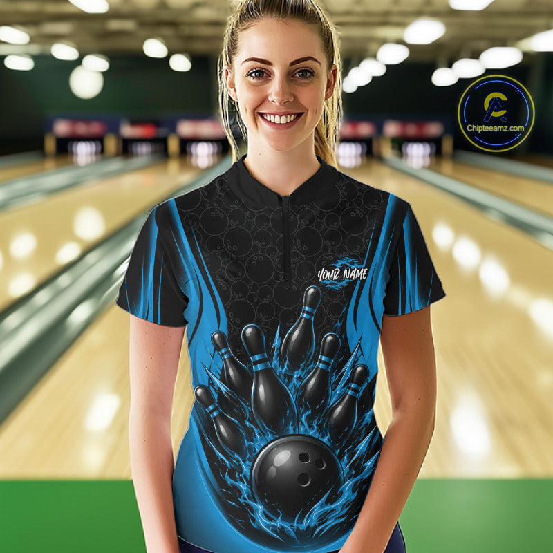 A woman stands in a bowling alley wearing a short-sleeve bowling quarter zip shirt with a black and blue flame design, bowling pins, and a bowling ball graphic on the front. 