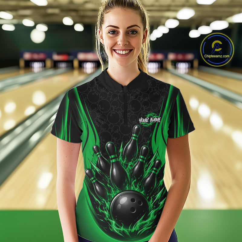 A woman stands in a bowling alley wearing a custom bowling quarter zip  jersey with a black and green flame design, bowling pins, and a bowling ball graphic on the front. 