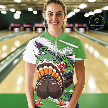 Front view of a happy female model in a bowling alley wearing a green custom quarter zip bowling shirt with a funny graffiti turkey holding a bowling ball, paint splatter accents, and a personalized name.