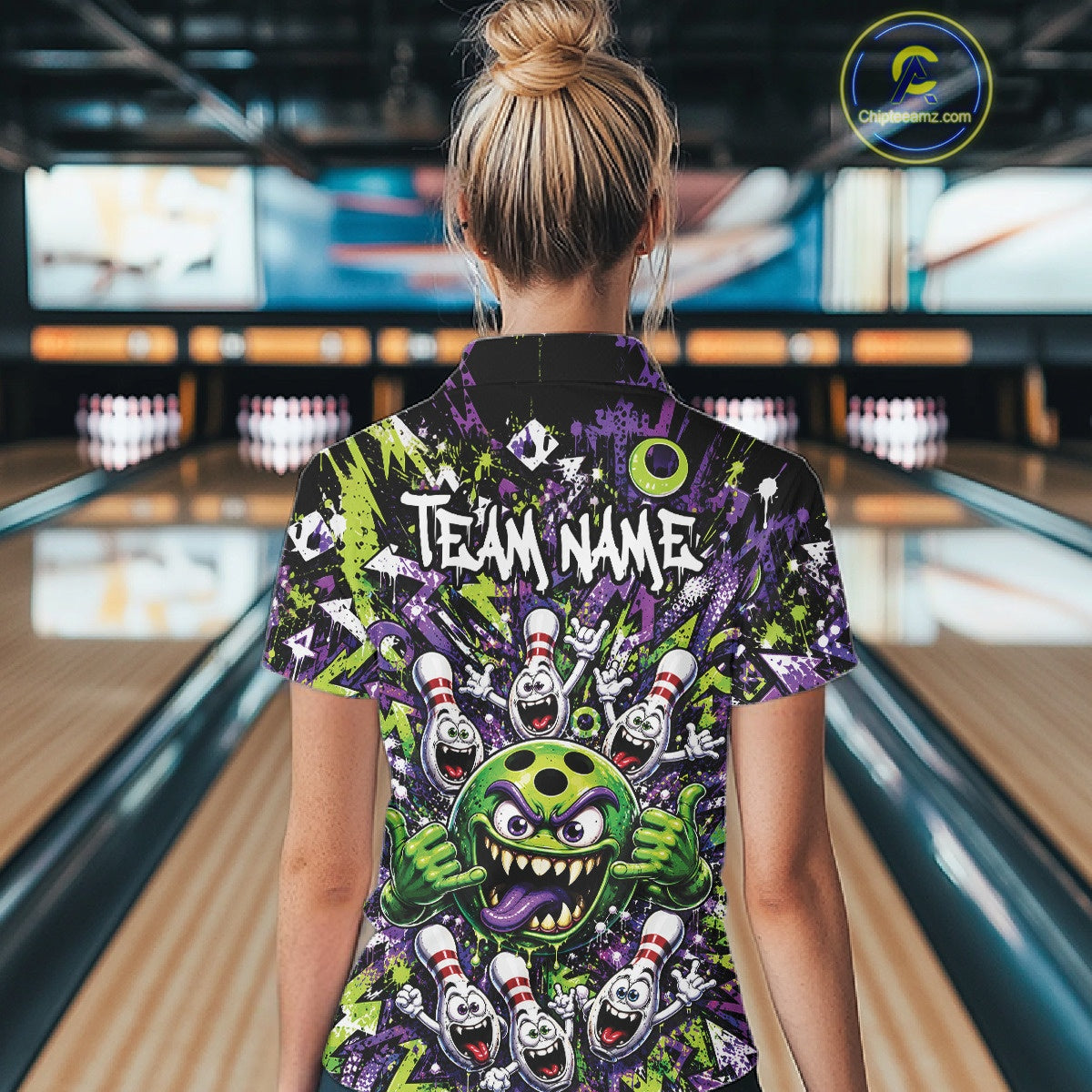 Back view of a female model in a bowling lane wearing a green and purple bowling quarter-zip shirt showcasing a mischievous green monster bowling ball with sharp teeth, a purple tongue, playful hand gesture, and cheerful cartoon bowling pins.