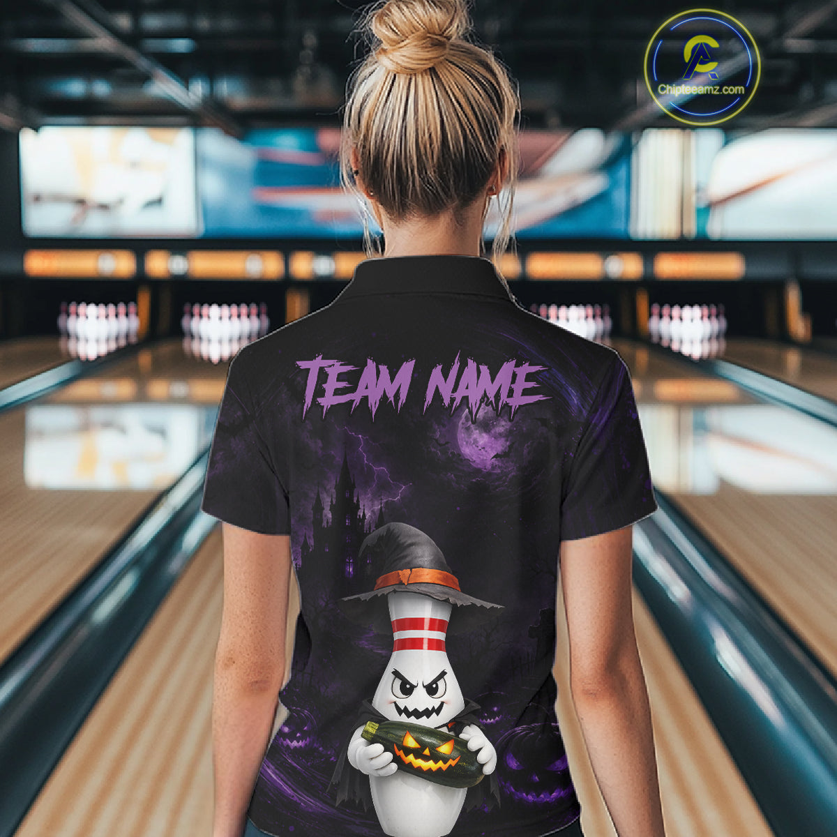 A woman viewed from behind wearing a black short-sleeve polo shirt with a Halloween-themed graphic print. The shirt features purple 'TEAM NAME' text and a cartoon ghost wearing a witch hat holding a pumpkin.