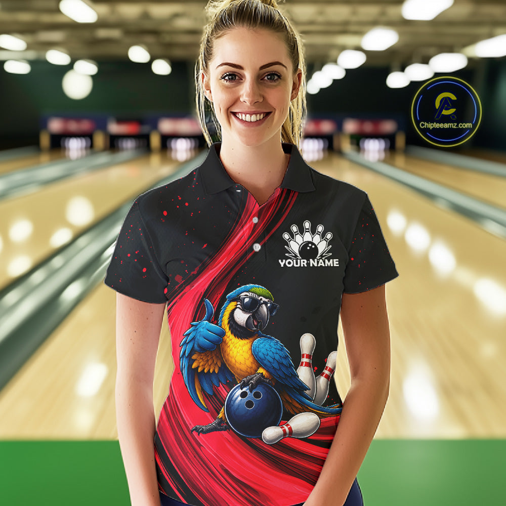 A woman stands in a bowling alley wearing a black short-sleeve polo shirt with a red graphic design, a blue and yellow parrot, bowling pins, and a blue bowling ball. The shirt also features a white bowling pin graphic and the text 'YOUR NAME' on the chest.