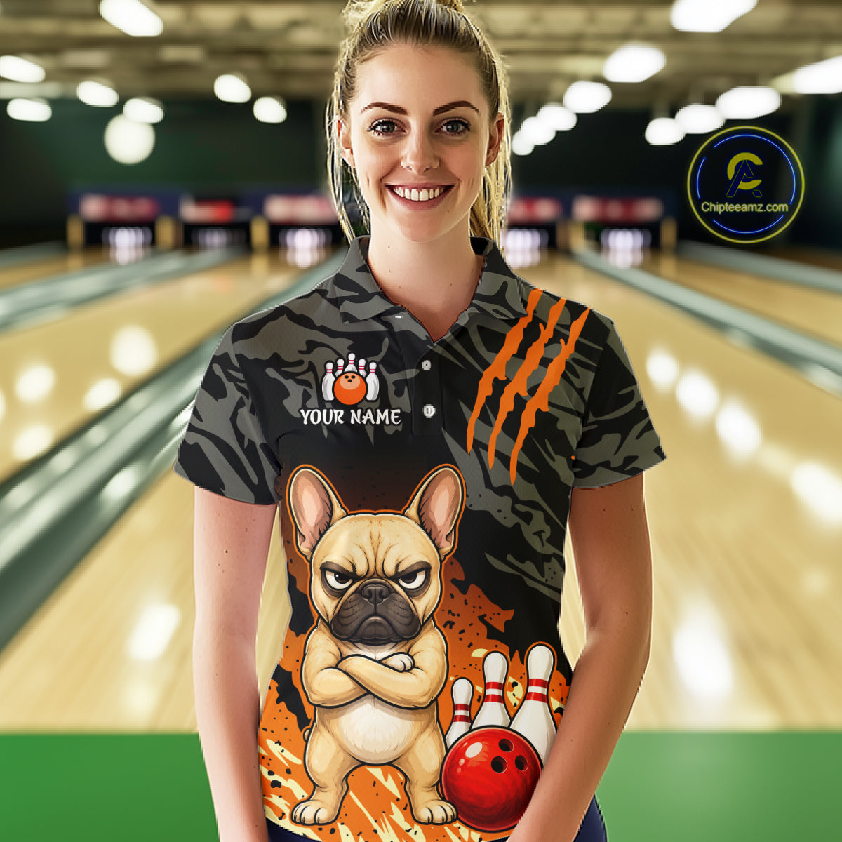 A woman stands in a bowling alley wearing a short-sleeve polo shirt with a dark camouflage pattern, orange claw-stripe graphics, a cartoon French bulldog, bowling pins, and a red bowling ball. The shirt also has a customizable name patch on the chest.