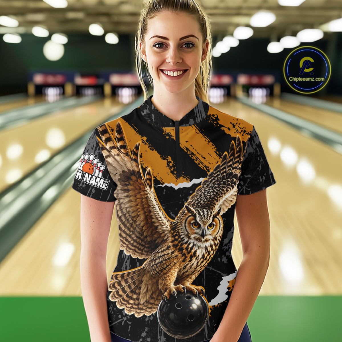 A woman standing in a bowling alley wearing a short-sleeve bowling shirt with a large owl graphic and bowling-themed text. The background shows blurred bowling lanes with lights and pins.