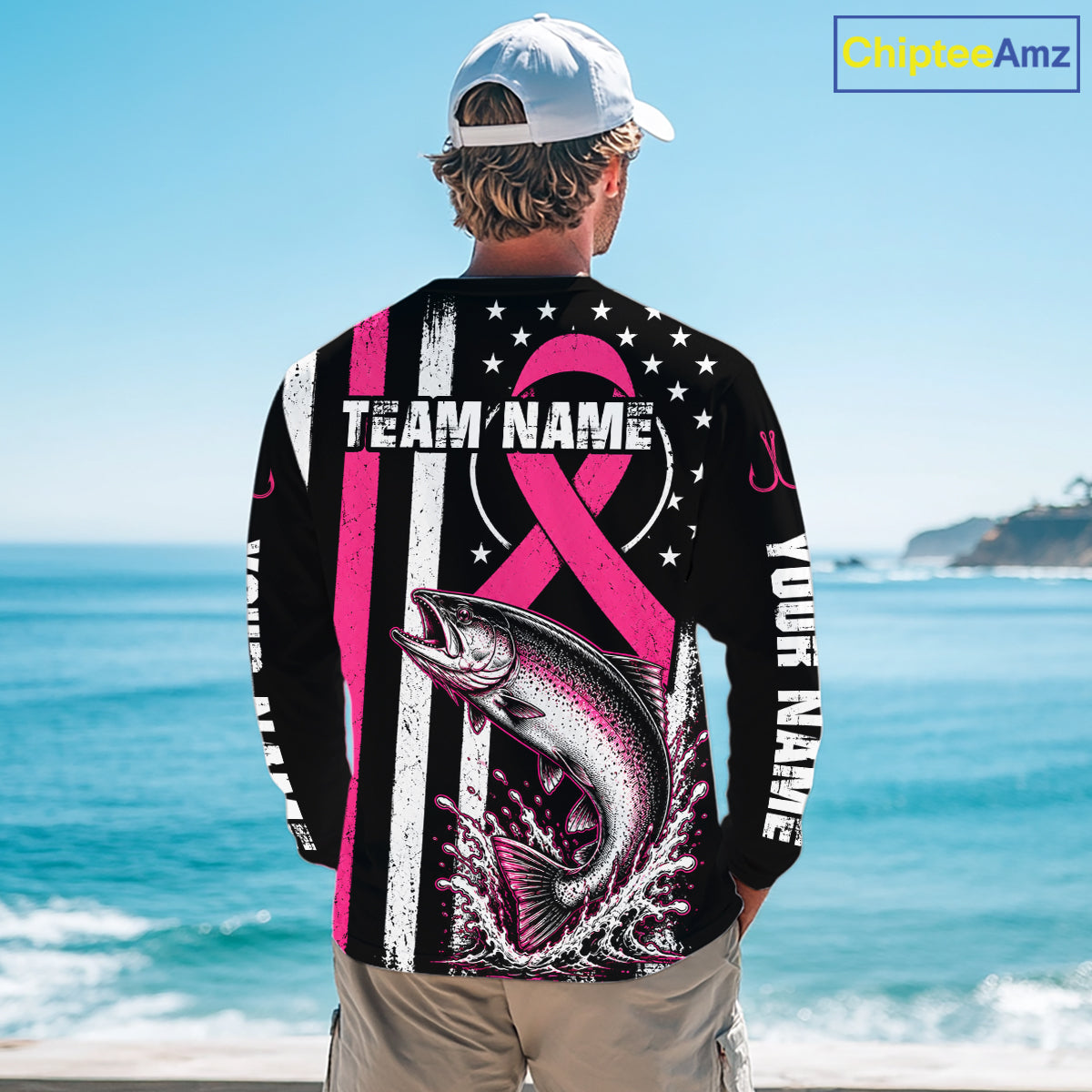 A man stands on a beach facing away from the camera, wearing a black long-sleeve shirt with a large pink and white graphic design featuring a fish, stars, stripes, and customizable text. The background shows the blue ocean and sky.